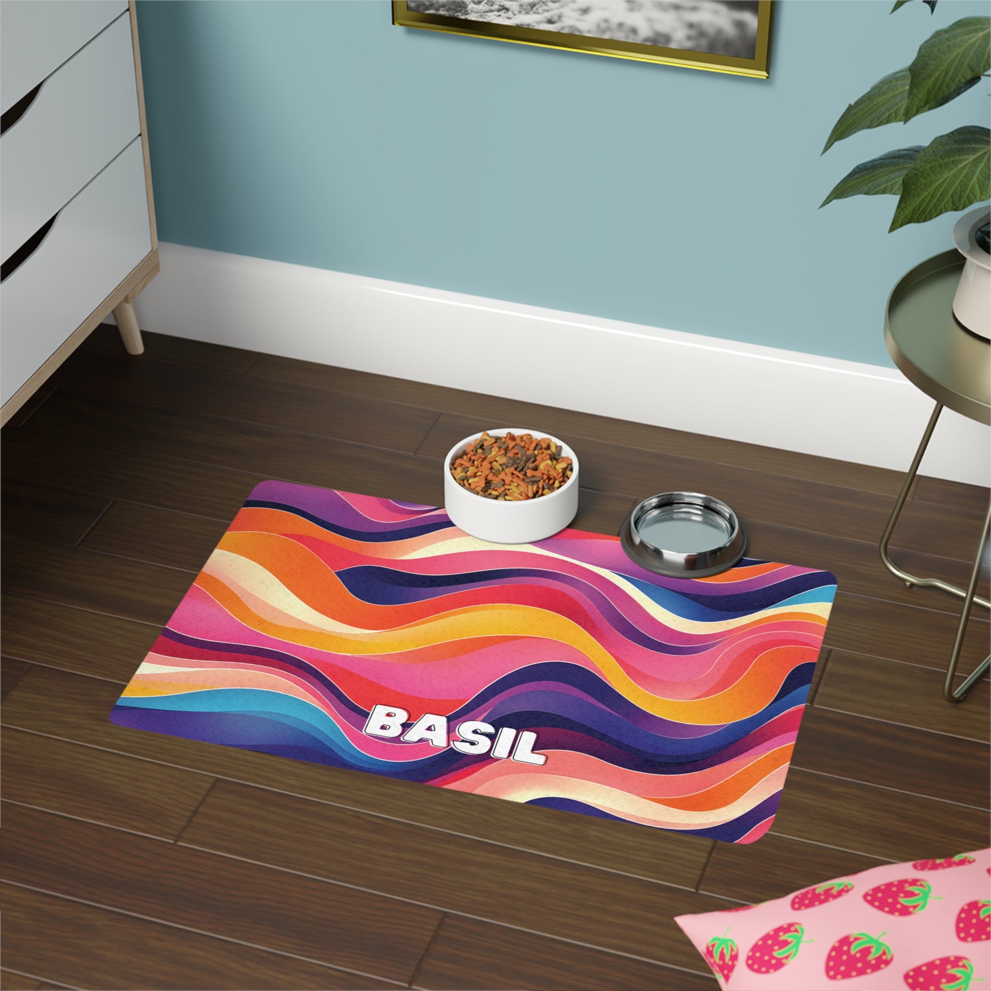 A colorful, African-inspired mat with wavy, abstract stripes in orange, pink, purple, and yellow tones sits on a dark wooden floor. “BASIL” is written in bold white letters. A bowl of pet food and water dish rest on the mat near a light blue wall.