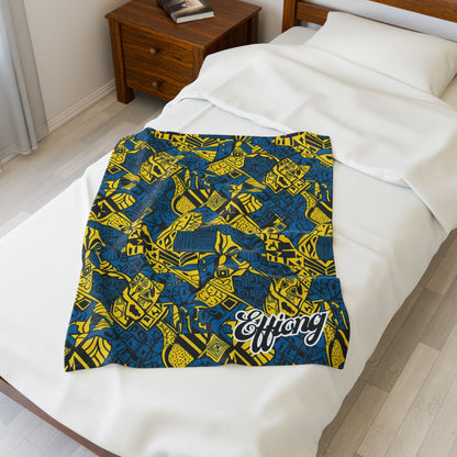 A colorful African-inspired blanket with bold blue and yellow geometric patterns and the word Effiong in white script is spread over a neatly made bed beside a wooden nightstand in a bright, minimalist bedroom.