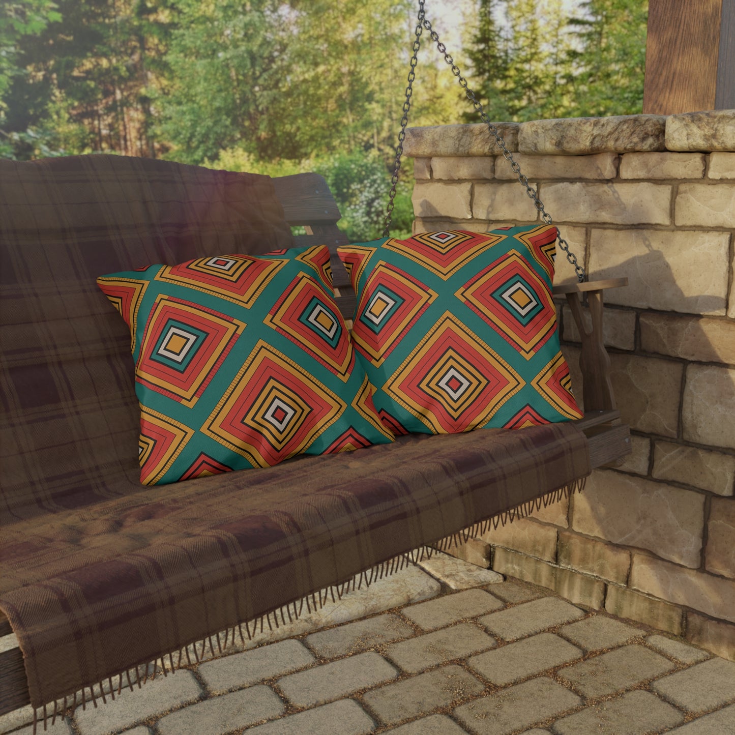 A wooden porch swing with a plaid blanket and two outdoor pillows in colorful, geometric patterns sits on a stone-paved patio. The swing is next to a stone wall, with lush green trees in the sunlit background.