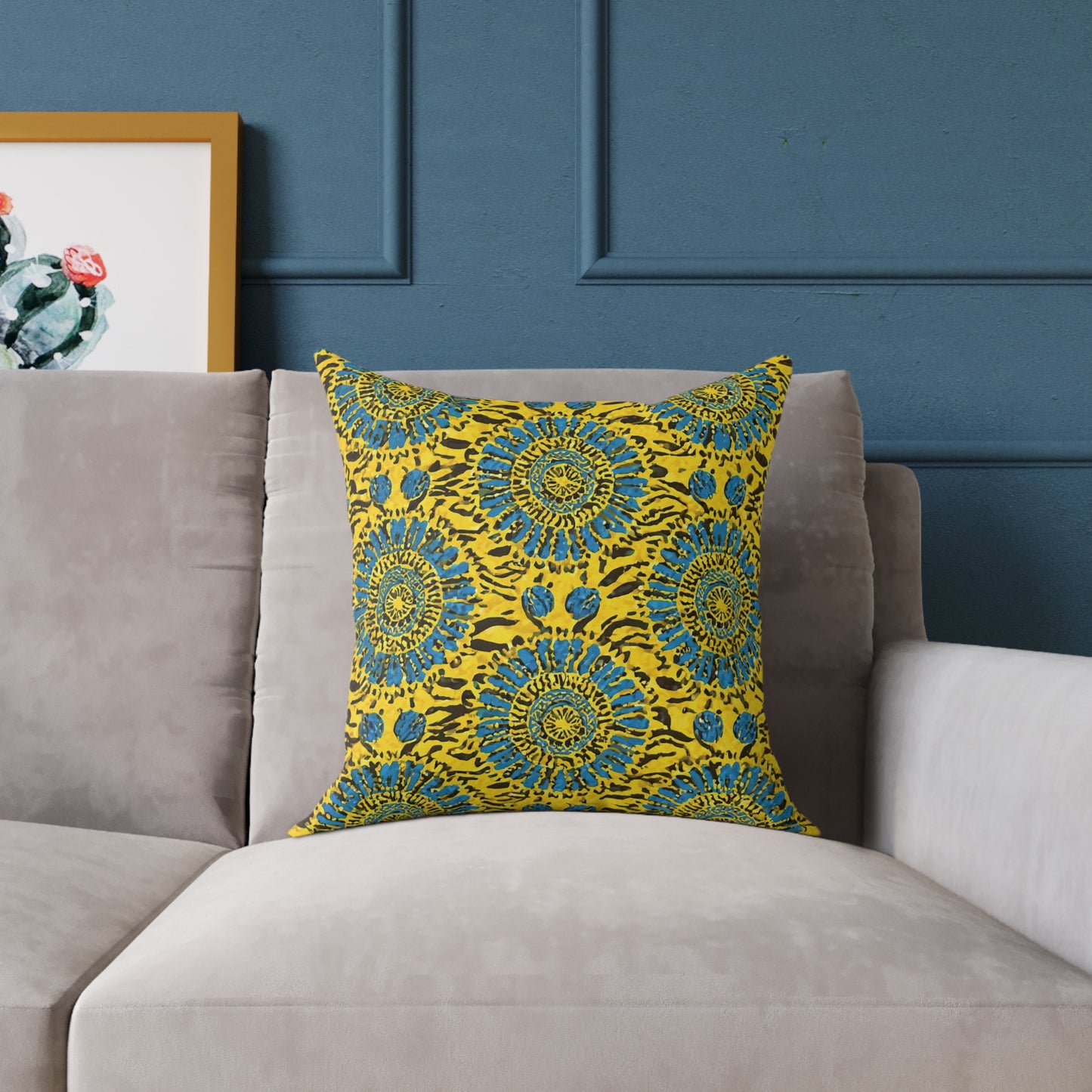 A vibrant throw pillow with a bright yellow cover and an intricate blue and black floral mandala pattern adds a touch of colorful home decor to the light gray sofa, set against a dark blue paneled wall with a framed cactus print peeking through.