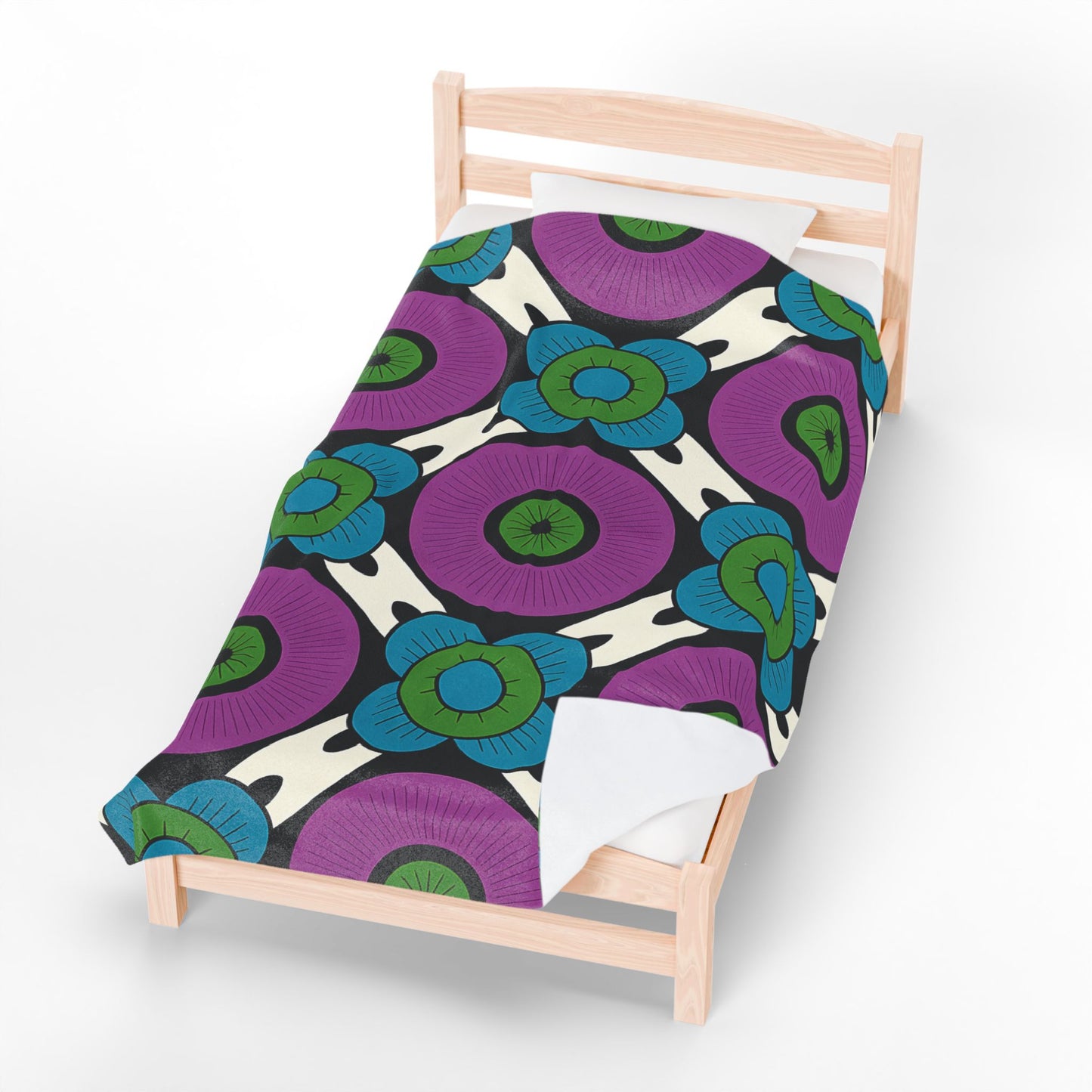 A light wood single bed with a custom velveteen blanket featuring a bold floral pattern: large purple circles with green centers, blue outer petals, and dark lines on an off-white background. Perfect as a unique gift, set on a plain white floor.