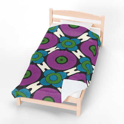 A light wood single bed with a custom velveteen blanket featuring a bold floral pattern: large purple circles with green centers, blue outer petals, and dark lines on an off-white background. Perfect as a unique gift, set on a plain white floor.