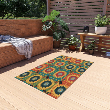 A colorful African-inspired outdoor rug with red, yellow, and green starburst patterns lays on a light wooden deck. Its no-slip backing ensures safety, while wicker furniture and potted plants create a cozy, modern outdoor seating area.