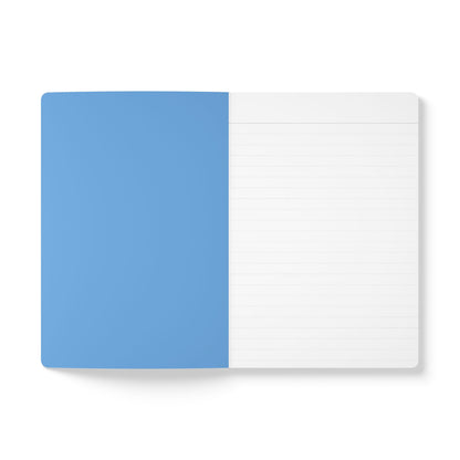 An open creative journal lies flat on a white surface. The left page is solid light blue and blank, while the right page is white with faint gray horizontal lines. The notebook has rounded corners and a minimal, clean design.