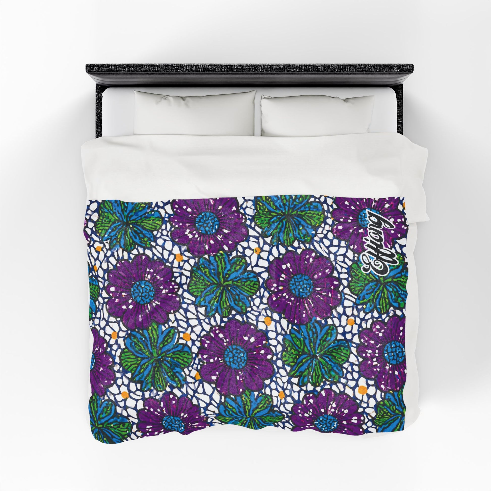 A bed with a white duvet featuring a vibrant pattern of large purple flowers, green leaves, blue accents, and small orange dots. The background has a mosaic-like design, and “Akong” is written in cursive—an African-inspired personalized blanket.