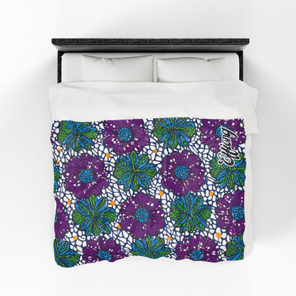 A bed with a white duvet featuring a vibrant pattern of large purple flowers, green leaves, blue accents, and small orange dots. The background has a mosaic-like design, and “Akong” is written in cursive—an African-inspired personalized blanket.
