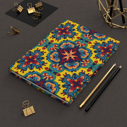 A colorful hardcover journal with an ornate, symmetrical floral pattern in blue, red, and yellow lies on a textured brown surface. Nearby are a gold pen, black pencil, gold binder clips, and a geometric gold wire decor piece.