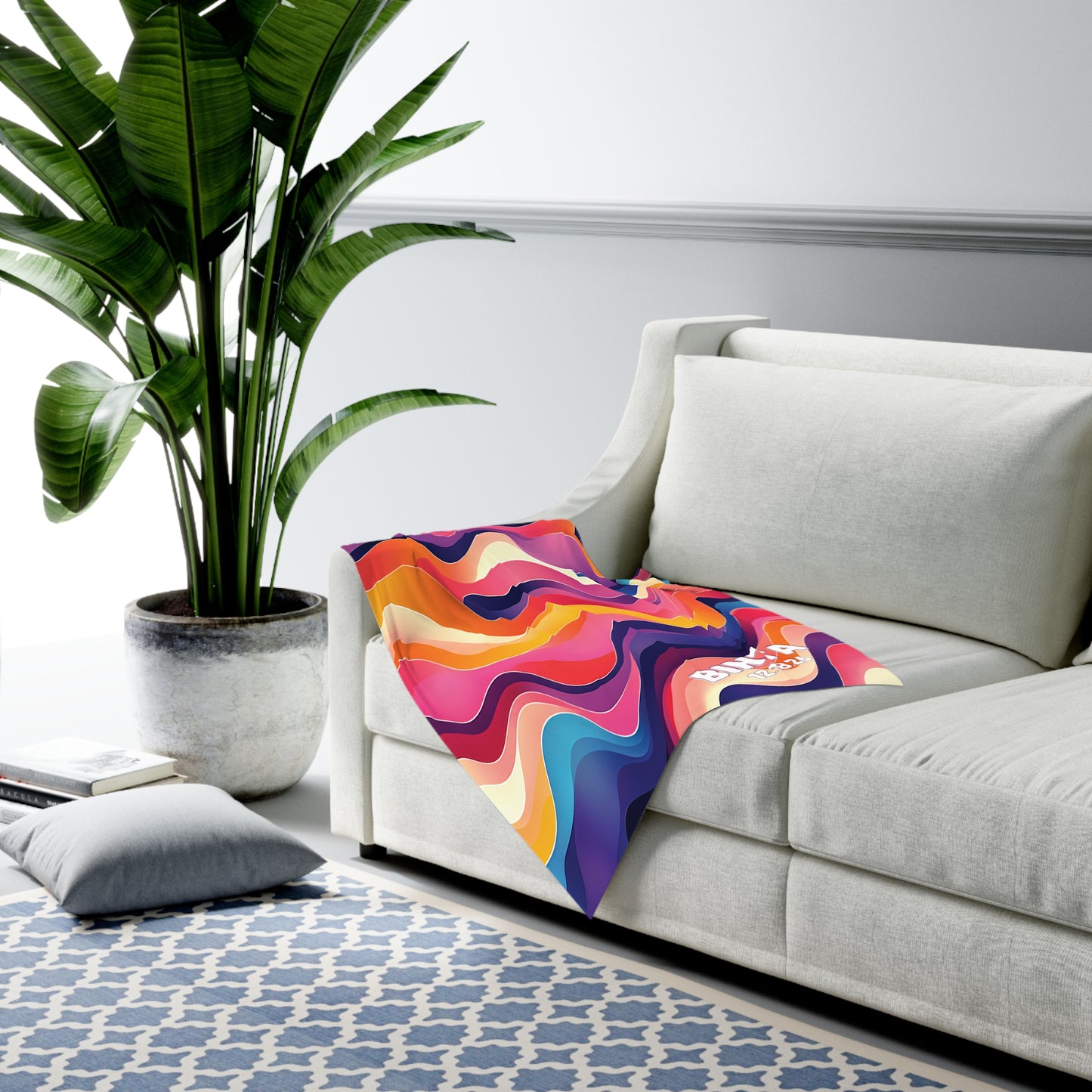 A modern living room features a large green potted plant, a light-colored sofa with an African-inspired decor touch—a colorful, wavy-patterned custom swaddle blanket draped over the arm—and a blue-and-white geometric rug. A closed book and a gray cushion lie on the floor.