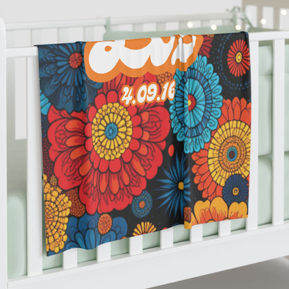 A colorful personalized baby blanket with large, bold floral patterns in red, yellow, orange, and blue drapes over a white crib rail. The blanket features bold white and orange text, including the date 4.09.16. String lights glow softly in the background.