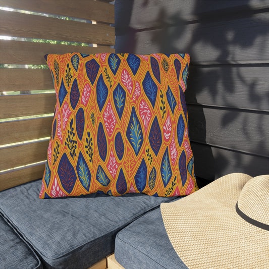 A colorful, African-inspired pillow with a bold leaf pattern in orange, pink, blue, and navy rests on a gray outdoor cushion. Sunlight and shadows play across a wooden privacy screen and dark wall, with a straw hat lying nearby.