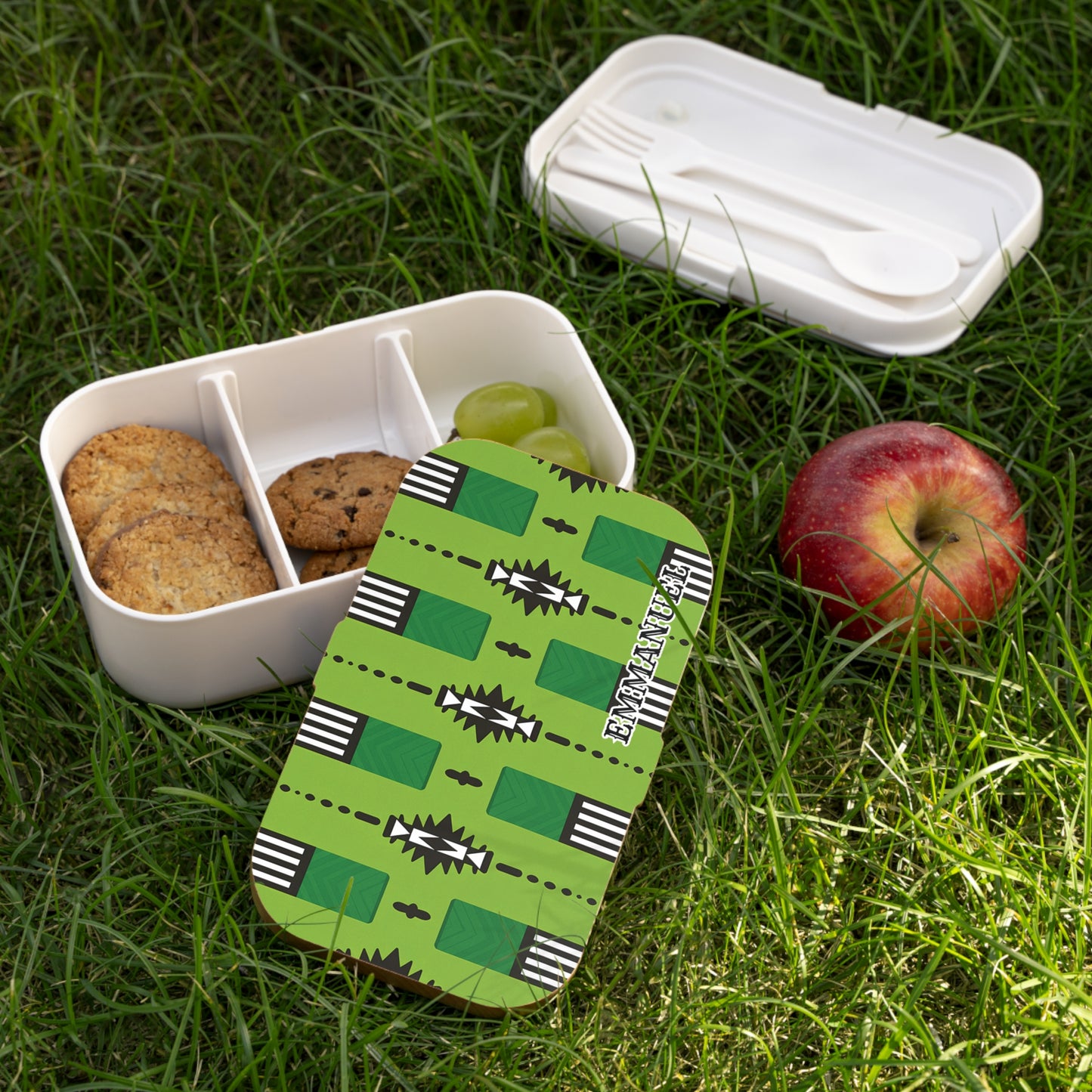 An African-inspired bento lunch box with cookies and grapes sits on grass. Its lid, bearing a green geometric pattern and “EKINATAH,” is removed. Beside it are a BPA-free utensil box with cutlery and a red apple.