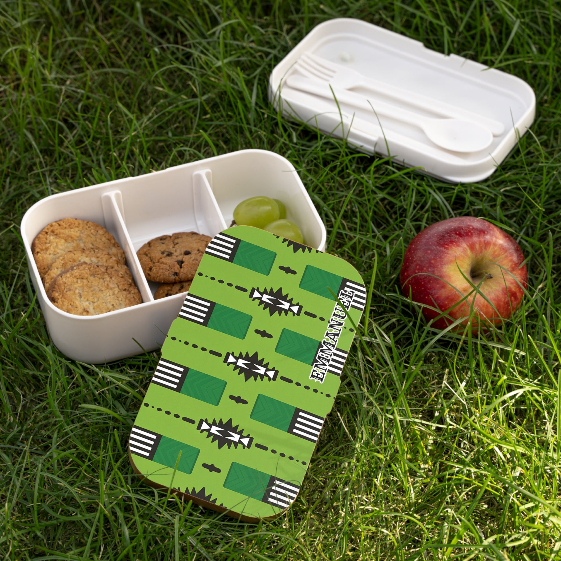 An African-inspired bento lunch box with cookies and grapes sits on grass. Its lid, bearing a green geometric pattern and “EKINATAH,” is removed. Beside it are a BPA-free utensil box with cutlery and a red apple.