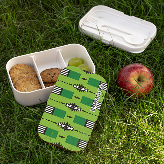 An African-inspired bento lunch box with cookies and grapes sits on grass. Its lid, bearing a green geometric pattern and “EKINATAH,” is removed. Beside it are a BPA-free utensil box with cutlery and a red apple.