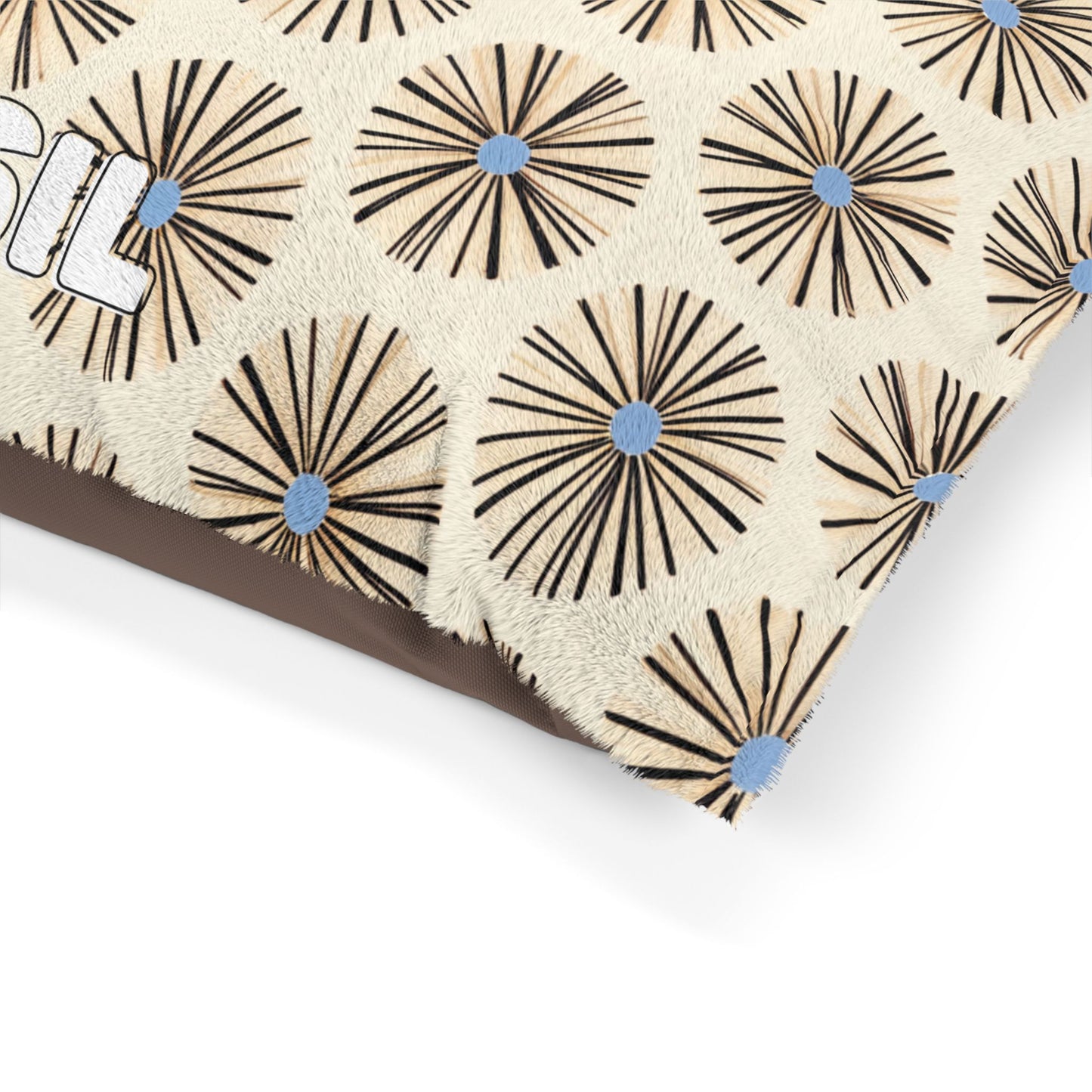 A close-up of an African-inspired beige cushion with a pattern of radiating black lines and blue dots, resembling abstract flowers or sunbursts. Part of the word SIL is visible in white outlined letters on the left. The cushion rests on a brown surface.