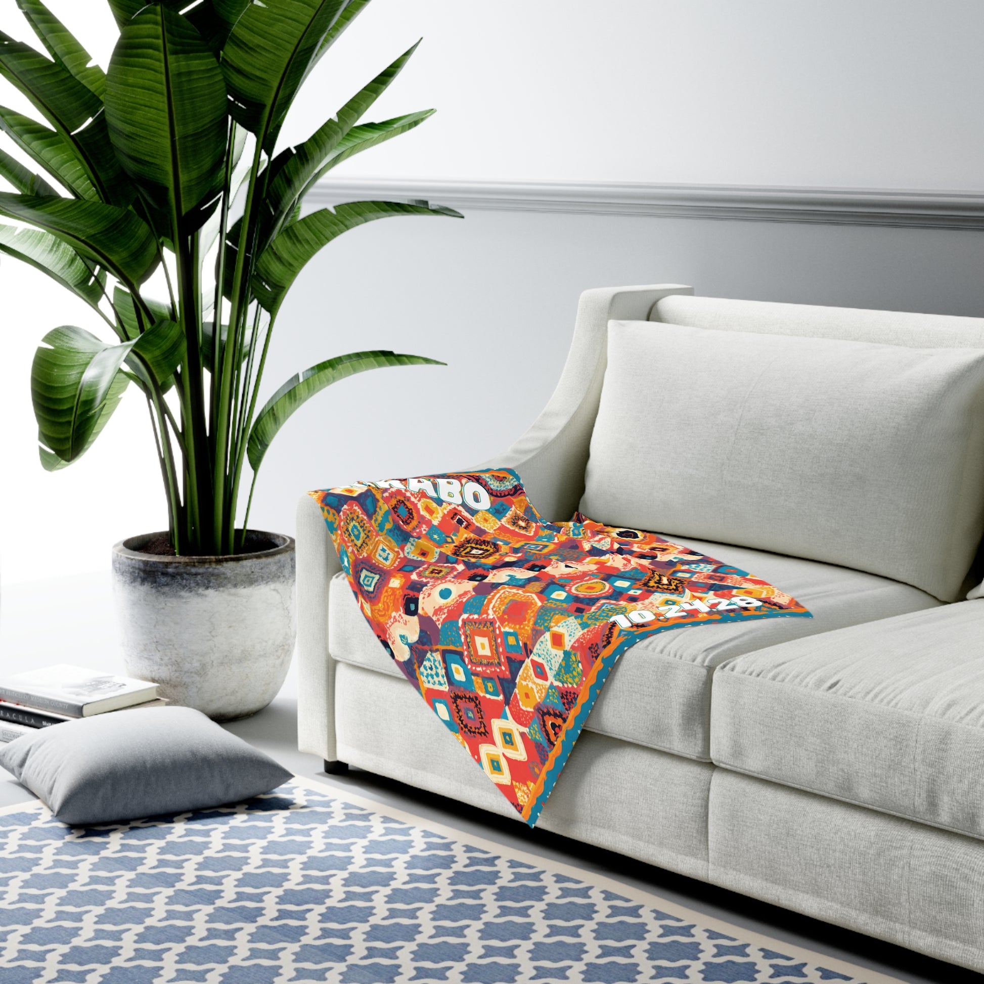 A white modern sofa with a colorful geometric-patterned, African-inspired decor blanket draped over the arm sits beside a large potted plant. A blue and white patterned rug and a gray floor cushion complete this bright, minimal living room.