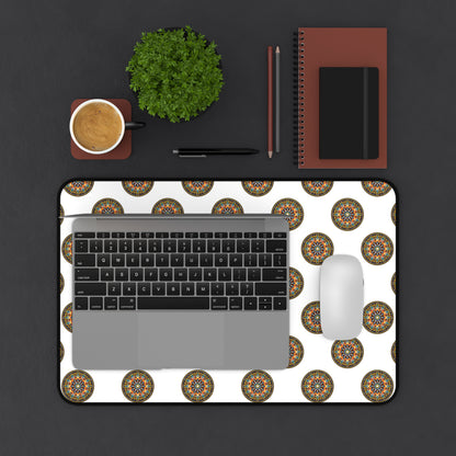 A top view of a desk with a laptop, mouse, and coffee on a colorful workspace accessory—a white Neoprene desk mat with circular mandala patterns. Nearby are a green potted plant, two pencils, a black pen, and stacked brown notebooks on a dark surface.
