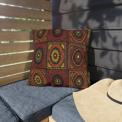 A decorative weatherproof pillow with a bold geometric and tribal-style pattern in red, yellow, and green rests on a blue outdoor cushion. The background features wooden slats and a straw hat nearby, casting a shadow on the cushion.