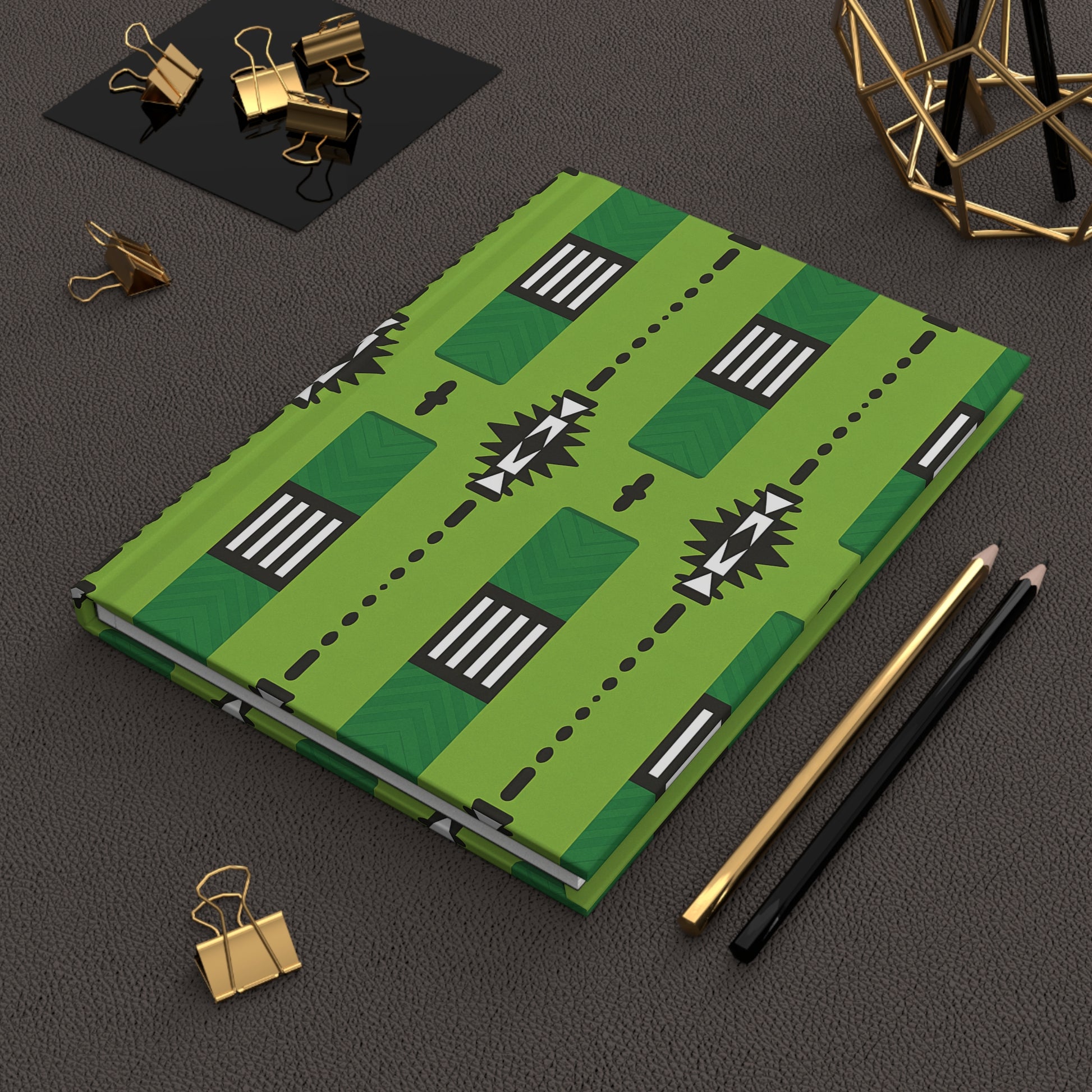 An African-inspired notebook with geometric black and white patterns lies on a textured dark surface beside a gold binder clip, two pencils (one gold, one black), a black tray with more gold clips, and a metallic wire decor object.