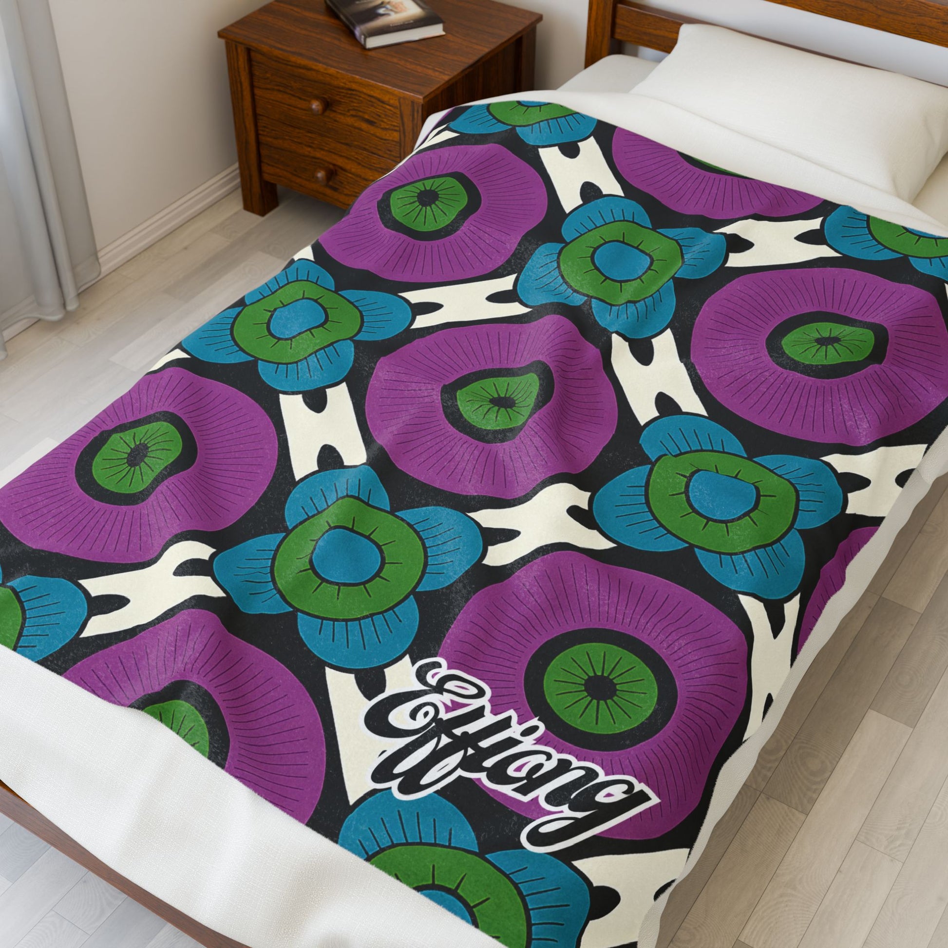 A bed with a vibrant velveteen blanket featuring large, stylized flowers in purple, green, and blue with dark centers on a white and black pattern. “Effang” is scripted at the bottom, making this an eye-catching African-inspired personalized blanket.
