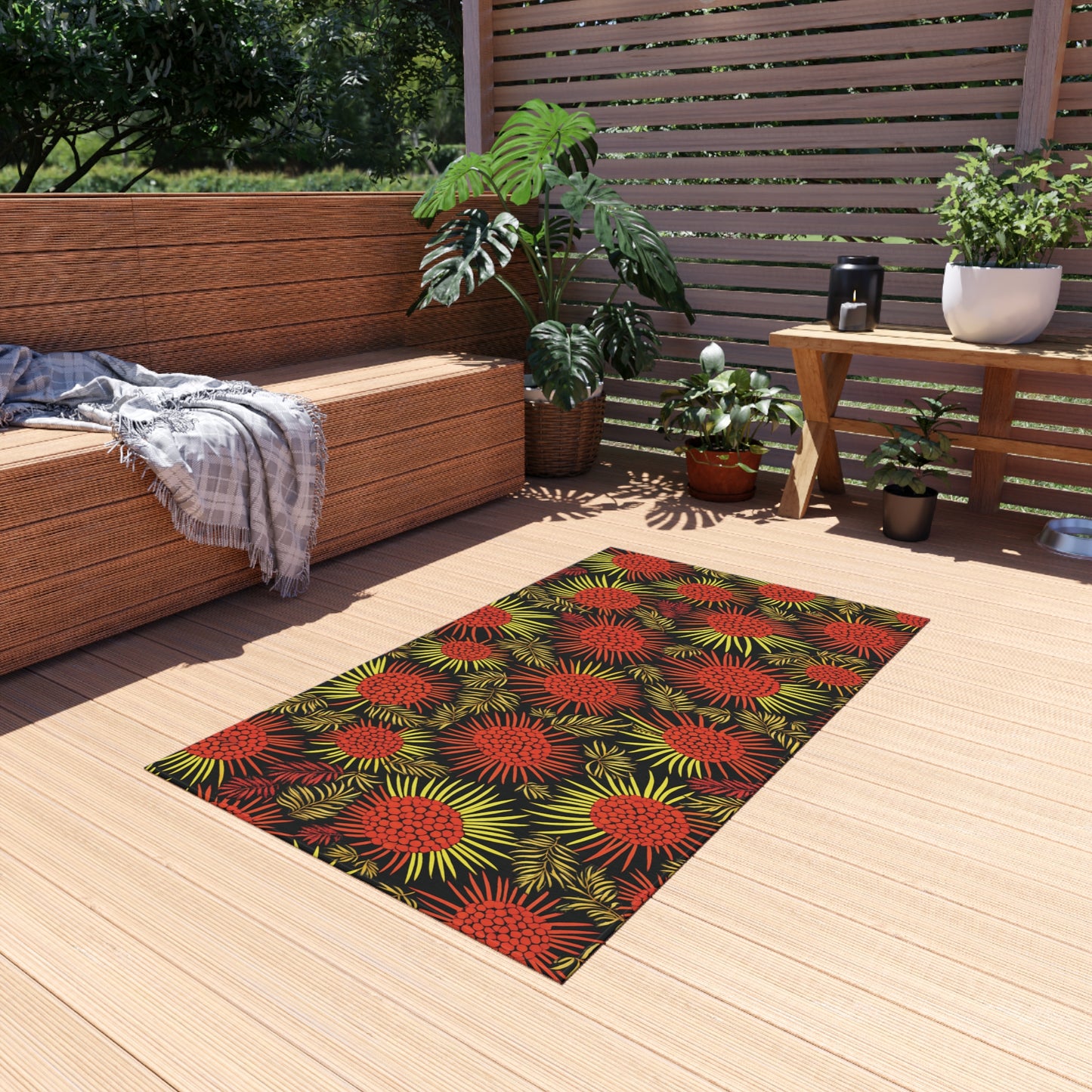 A modern patio features a tan wooden deck with an African-inspired rug patterned with red and yellow burst designs. There’s a wicker sofa with gray cushions and a plaid throw, lush green potted plants, and horizontal wood slat fencing.