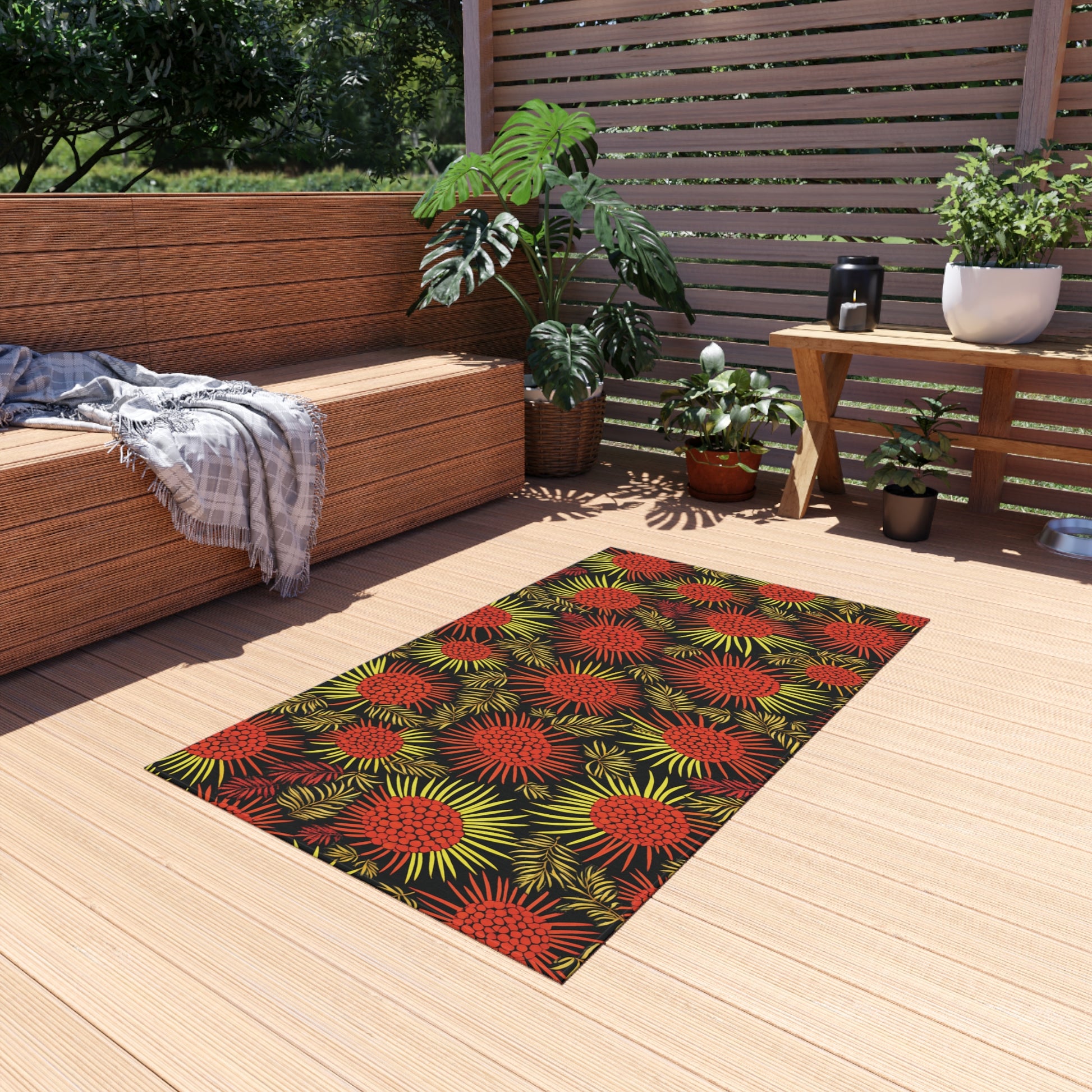 A modern patio features a tan wooden deck with an African-inspired rug patterned with red and yellow burst designs. There’s a wicker sofa with gray cushions and a plaid throw, lush green potted plants, and horizontal wood slat fencing.