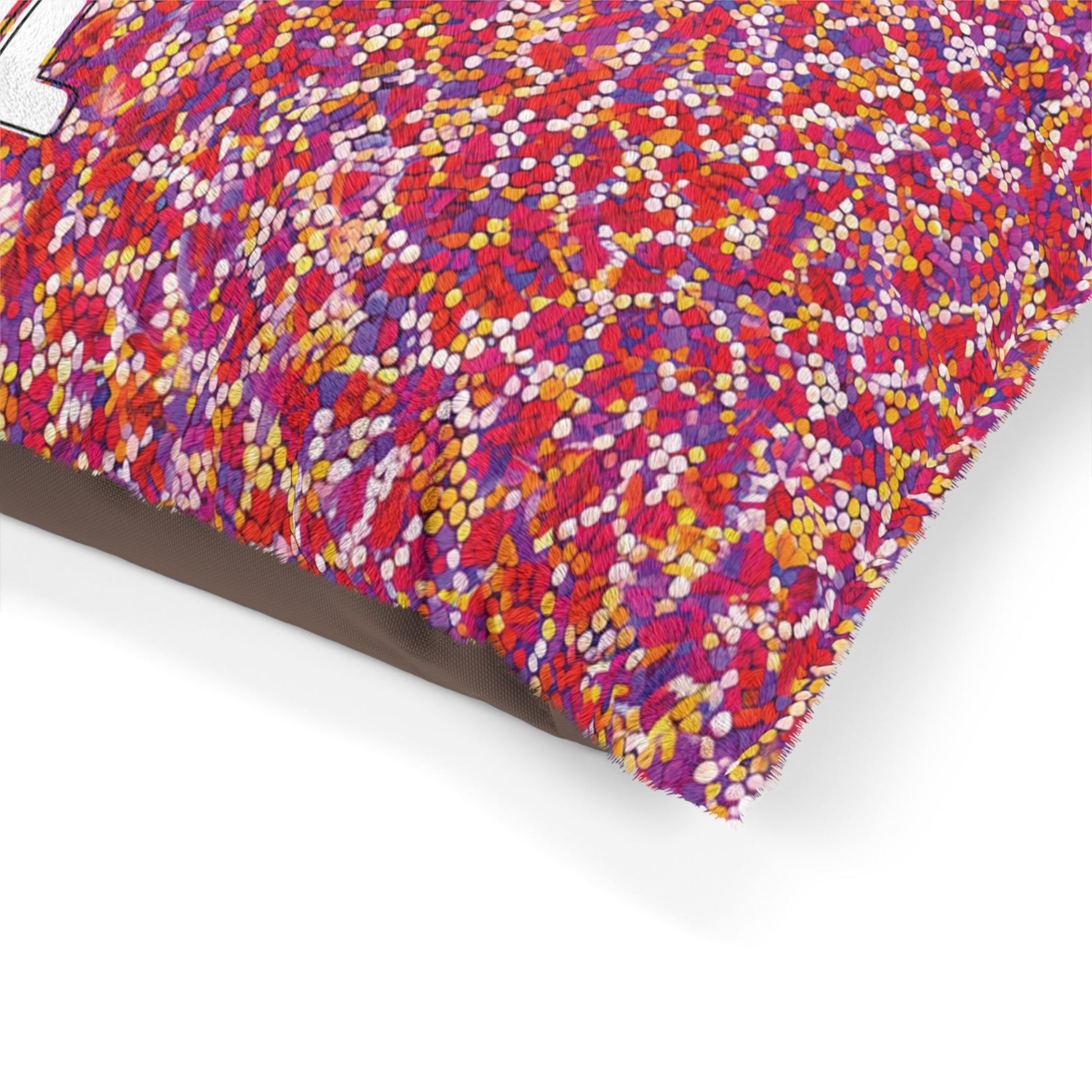 Close-up of a pillow corner with a textured fabric featuring a dense, colorful dotted pattern in red, orange, yellow, pink, and purple for a vibrant look. Durable construction ensures lasting quality as it rests on a plain surface with part of a white printed letter visible.