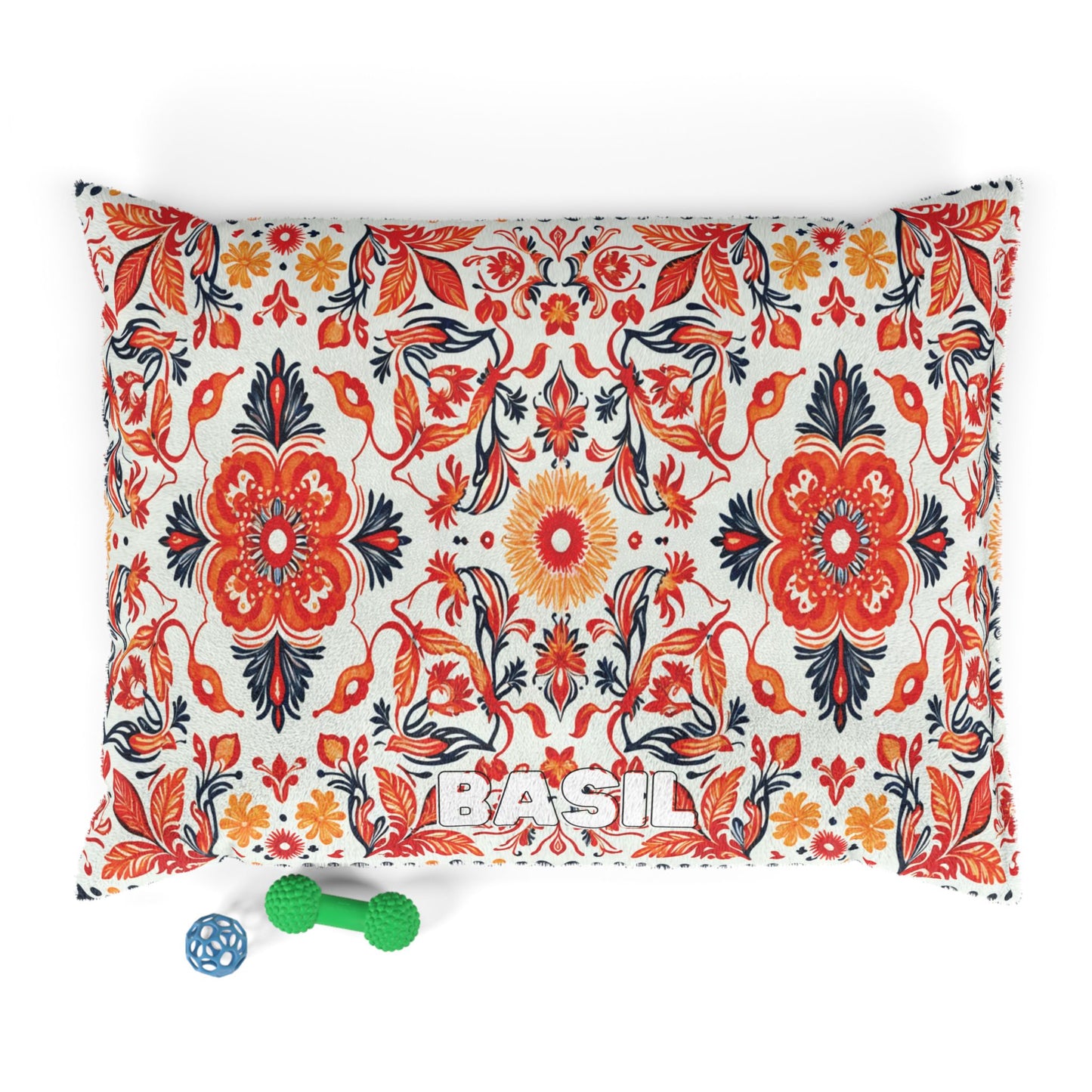 A rectangular pillow with a bold African-inspired red, orange, and black floral pattern on a white background. The word “BASIL” is embroidered at the bottom. In front are two green textured dog toys and one blue lattice ball toy.