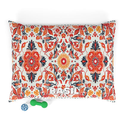 A rectangular pillow with a bold African-inspired red, orange, and black floral pattern on a white background. The word “BASIL” is embroidered at the bottom. In front are two green textured dog toys and one blue lattice ball toy.