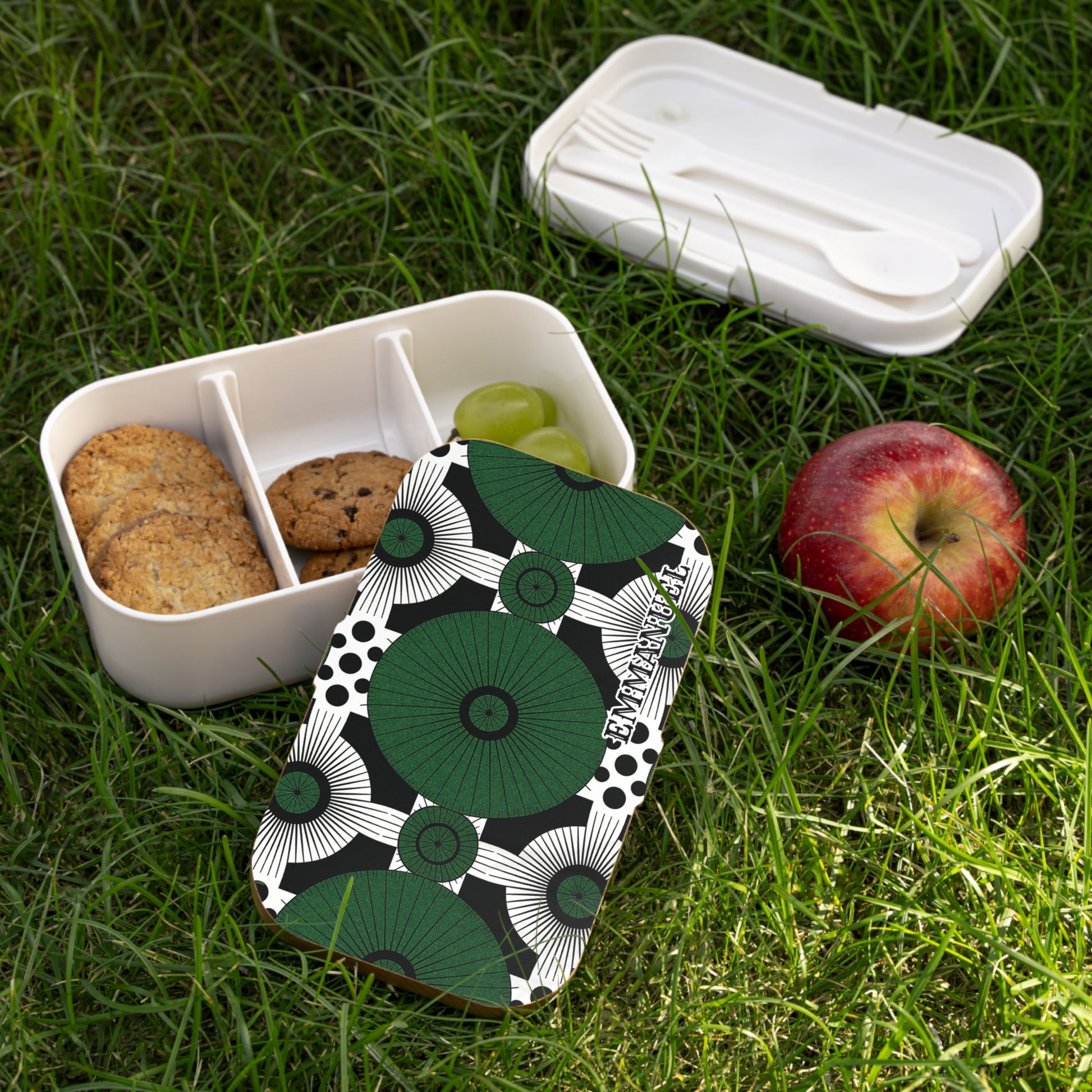 A white BPA-free lunchbox with a green and black geometric patterned lid sits open on grass. Inside, two oatmeal cookies, a chocolate chip cookie, and green grapes are arranged. Nearby, an apple and the open lid with white cutlery are visible.