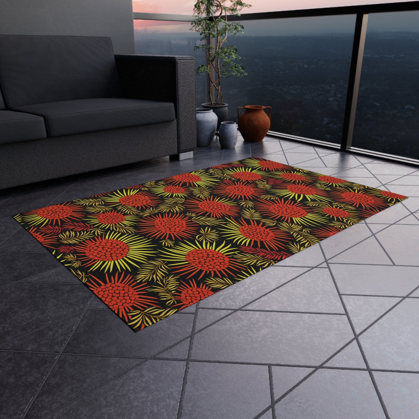 A modern balcony with a gray tiled floor features an African-inspired rectangular rug with a bold floral pattern in red, yellow, and green. A gray sofa, potted plants, and clay vases decorate the space. Cityscape and sunset shine through the glass railing.