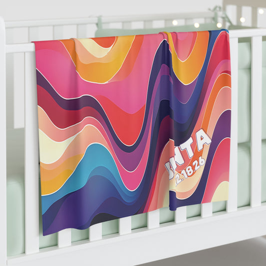 A colorful, African-inspired custom swaddle blanket with wavy stripes in pink, orange, yellow, blue, and purple hangs over a white crib. "INTA" and "12.18.26" appear in bold white letters with a playful font in the lower right corner.