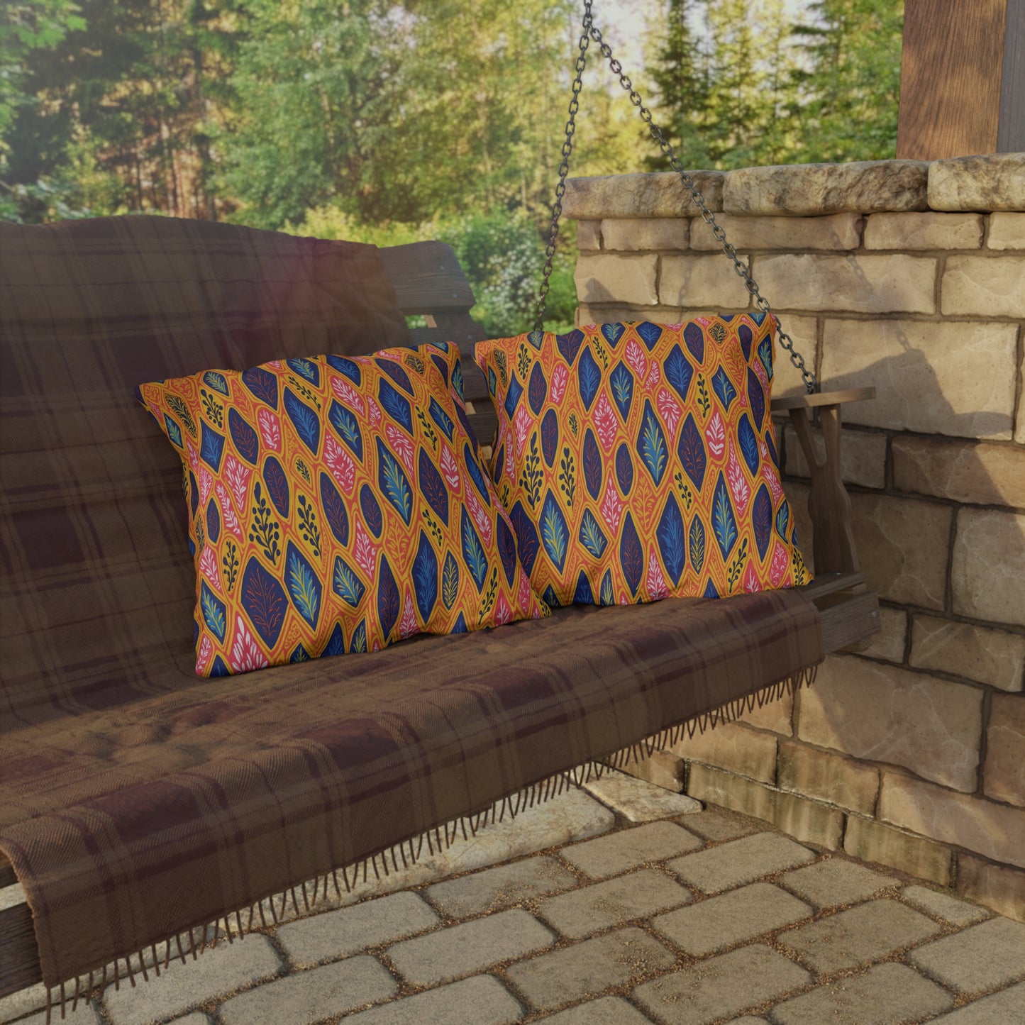 A wooden porch swing with a brown plaid blanket holds two colorful pillows featuring bold, African-inspired leaf patterns in orange, blue, and pink. The swing sits beside a stone wall, with greenery and trees visible in the background.