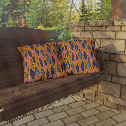 A wooden porch swing with a brown plaid blanket holds two colorful pillows featuring bold, African-inspired leaf patterns in orange, blue, and pink. The swing sits beside a stone wall, with greenery and trees visible in the background.