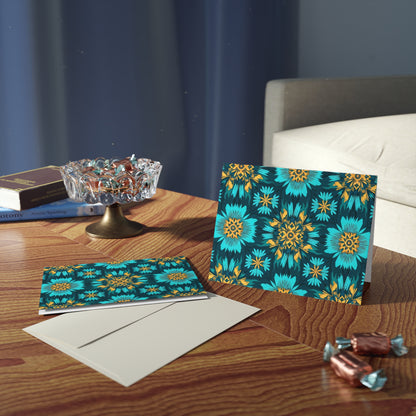 A wooden table holds two African-inspired greeting cards with vibrant teal and yellow floral patterns, premium stationery envelopes, an ornate glass bowl of wrapped candies, loose sweets, and stacked books beside a beige sofa and blue curtain.