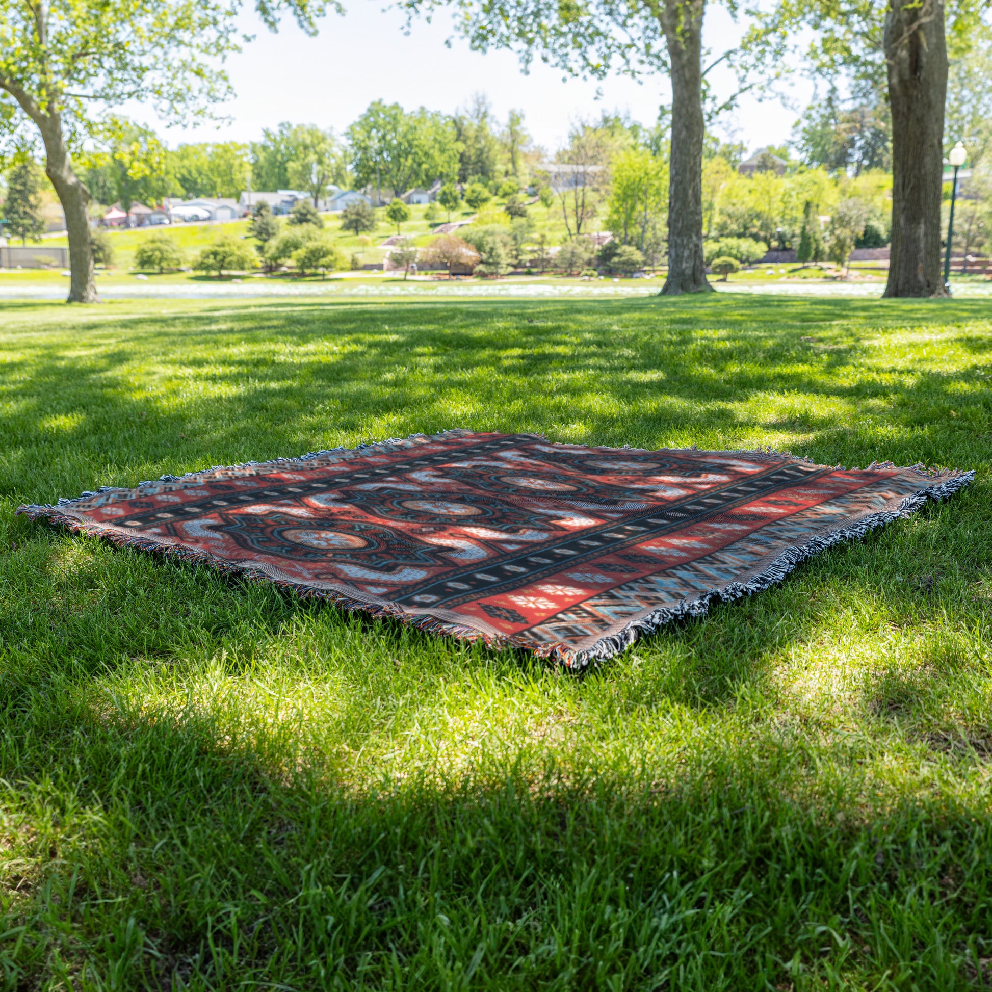 An African-inspired red and black patterned rug lies flat on green grass in a sunlit park. Tall trees cast dappled shade, with shrubs, open lawn, and distant parked cars visible under a clear sky.