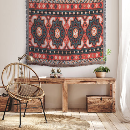 A cozy room features an African-inspired woven rattan chair, a wooden bench with books and a potted plant, and a large, colorful geometric tapestry with red, teal, and cream patterns. Sunlight filters through light curtains onto wooden floors.