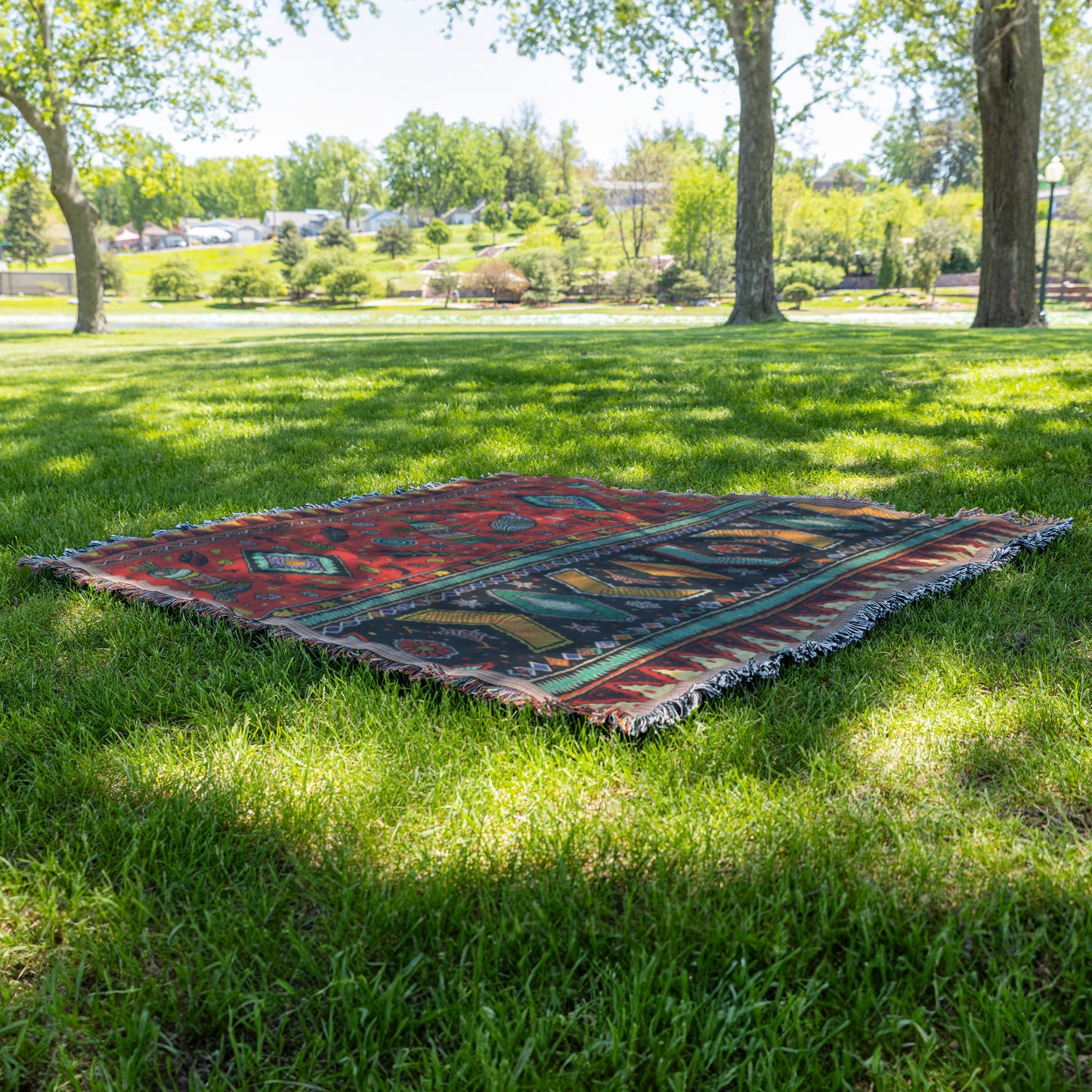 An African-inspired, colorful patterned blanket with red, blue, and green designs lays on green grass under the shade of large trees in a sunny park. Lush foliage, open grassy areas, and distant shrubs and trees are visible in the background.
