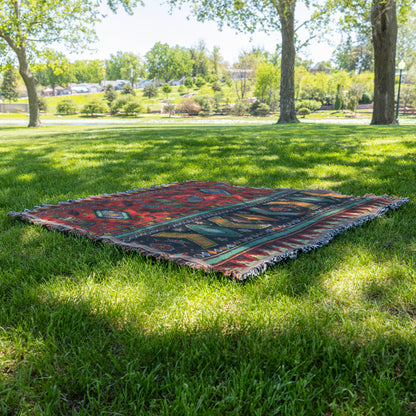 An African-inspired, colorful patterned blanket with red, blue, and green designs lays on green grass under the shade of large trees in a sunny park. Lush foliage, open grassy areas, and distant shrubs and trees are visible in the background.