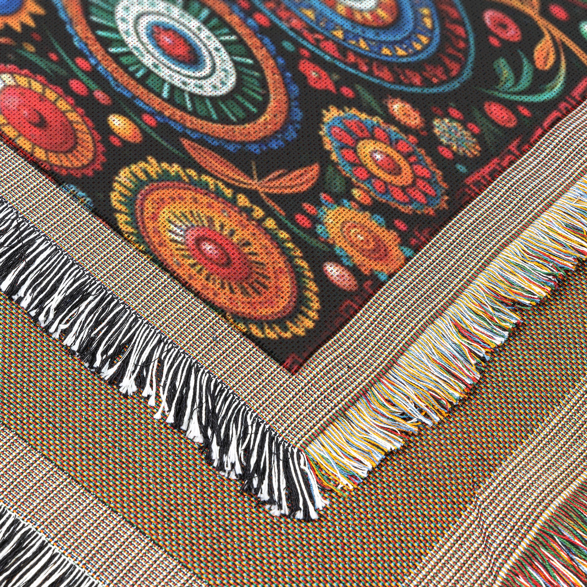 A close-up of a cozy weave textile featuring colorful, intricate circular patterns on a black background. The edge of the woven blanket has multicolored fringes, while an underlying layer shows beige and brown tones with a similar fringed border.