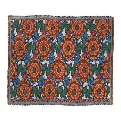 An African-inspired rectangular woven blanket with large red and orange flower patterns, green leaves, and white cloud-like shapes on a dark blue background. The blanket features fringed edges and a repeated, symmetrical floral design.