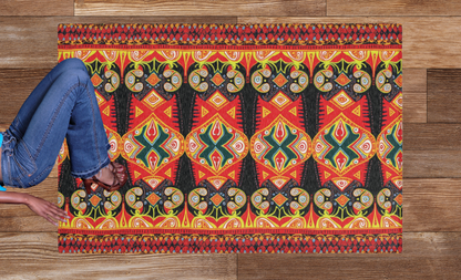 A person in blue jeans and brown sandals sits on a stylish colorful rug with red, yellow, green, and black geometric and tribal designs. This African design rug is placed on a wooden floor made up of planks in various shades of brown.