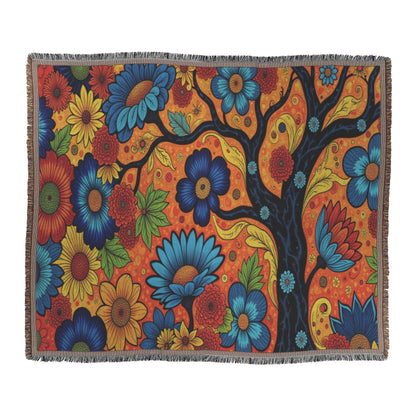 A vibrant throw blanket features a stylized tree with swirling black branches on an orange background reminiscent of West African print. Large, colorful flowers bloom across the scene, creating a lively, whimsical design with a fringed edge.