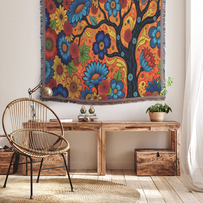 A cozy, boho-style interior features a rattan chair draped with a vibrant throw blanket, a wooden bench with books and a potted plant, and a tapestry depicting a black tree with colorful flowers on an orange background. Sunlight fills the room.