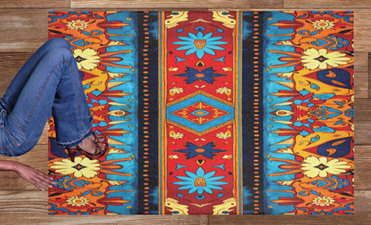A person in blue jeans and sandals sits on a wooden floor, beside a stylish colorful rug. The rug features symmetrical patterns in red, blue, yellow, and orange, with floral and geometric designs and a central diamond motif.