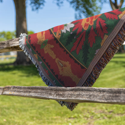 A colorful, African-inspired fringed blanket with red, green, and yellow floral patterns is draped over a rustic wooden fence. Green grass, leafy trees, and a blue sky in the background suggest a sunny outdoor setting.