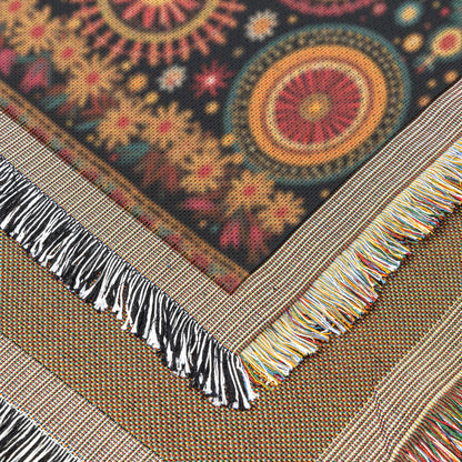 A close-up of a vibrant throw blanket with geometric, circular, and starburst patterns in red, gold, and teal on a black background. The woven blanket features fringed edges in white, black, and multicolored threads with overlapping corners shown.