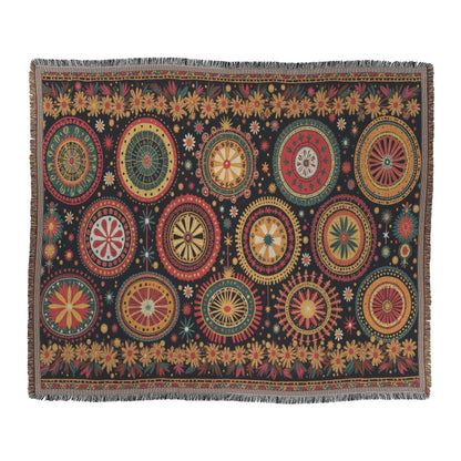 A rectangular textile with a black background features colorful, symmetrical circular patterns in red, yellow, green, and cream. Reminiscent of a West African print, this vibrant throw blanket includes smaller starburst and floral motifs and a short fringe on all edges.