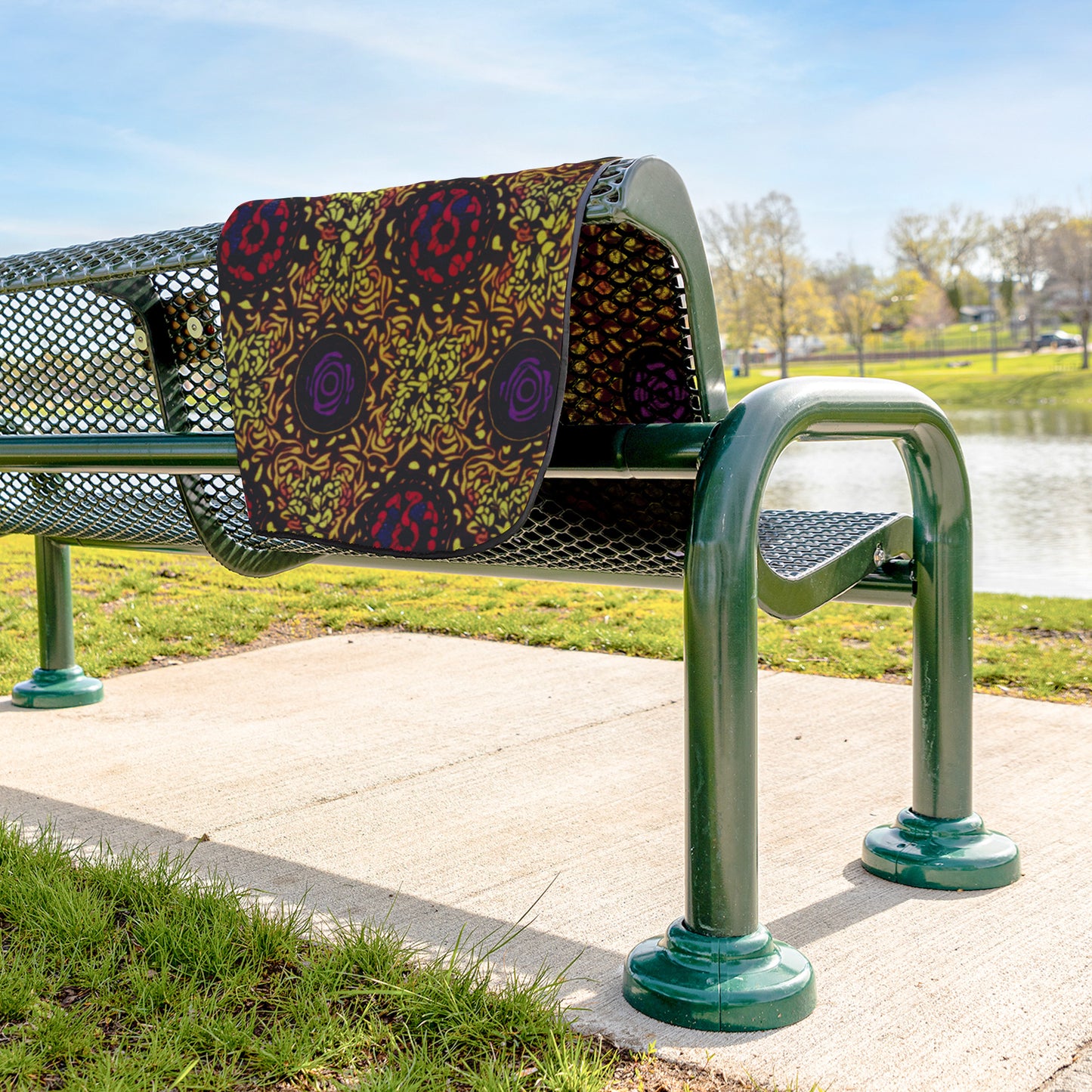 A green metal park bench sits on a paved sidewalk near grass and a pond. A water-resistant stadium blanket with bold, African-inspired patterns in red, purple, and yellow drapes over the bench. Trees and blue sky are visible in the background.