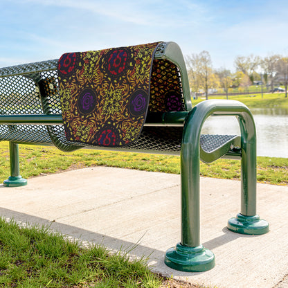 A green metal park bench sits on a paved sidewalk near grass and a pond. A water-resistant stadium blanket with bold, African-inspired patterns in red, purple, and yellow drapes over the bench. Trees and blue sky are visible in the background.