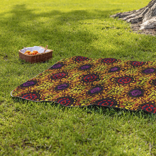 A picnic blanket with colorful, African-inspired patterns in red, yellow, and purple lies on green grass near a tree. To the left, a wicker basket with apples and a baguette sits on the grass in bright, natural sunlight.